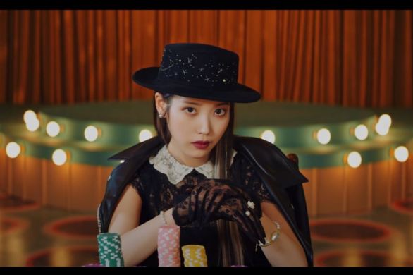 Screenshot of IU in the Coin music video. IU, dressed in classy clothes, at the end of the poker table with many poker chips in front of her. Behind her is an old-fashion stage (in which later in the video she sings on).