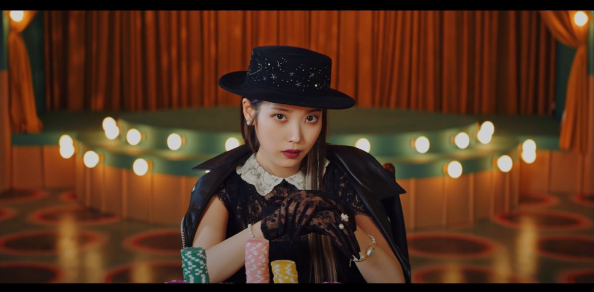 Screenshot of IU in the Coin music video. IU, dressed in classy clothes, at the end of the poker table with many poker chips in front of her. Behind her is an old-fashion stage (in which later in the video she sings on).